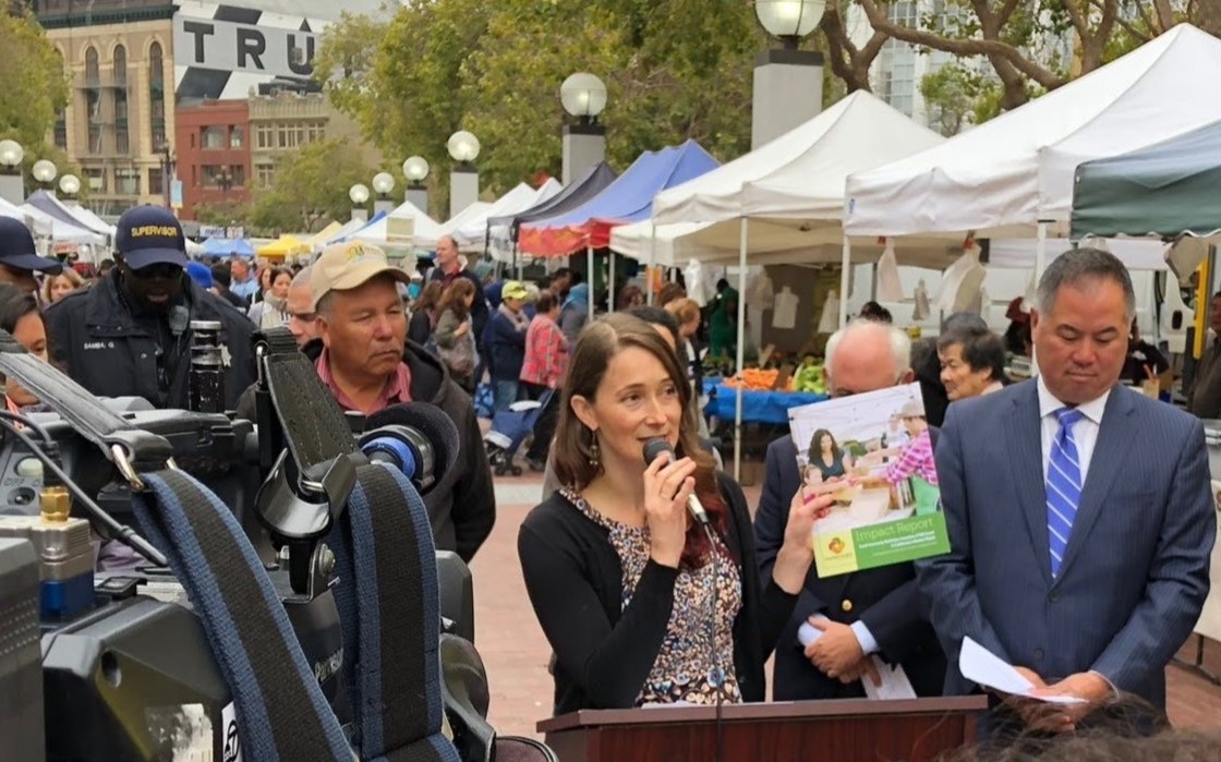 Photo at a press conference in a farmers' market. Carle is holding a report and speaking into a microphone.